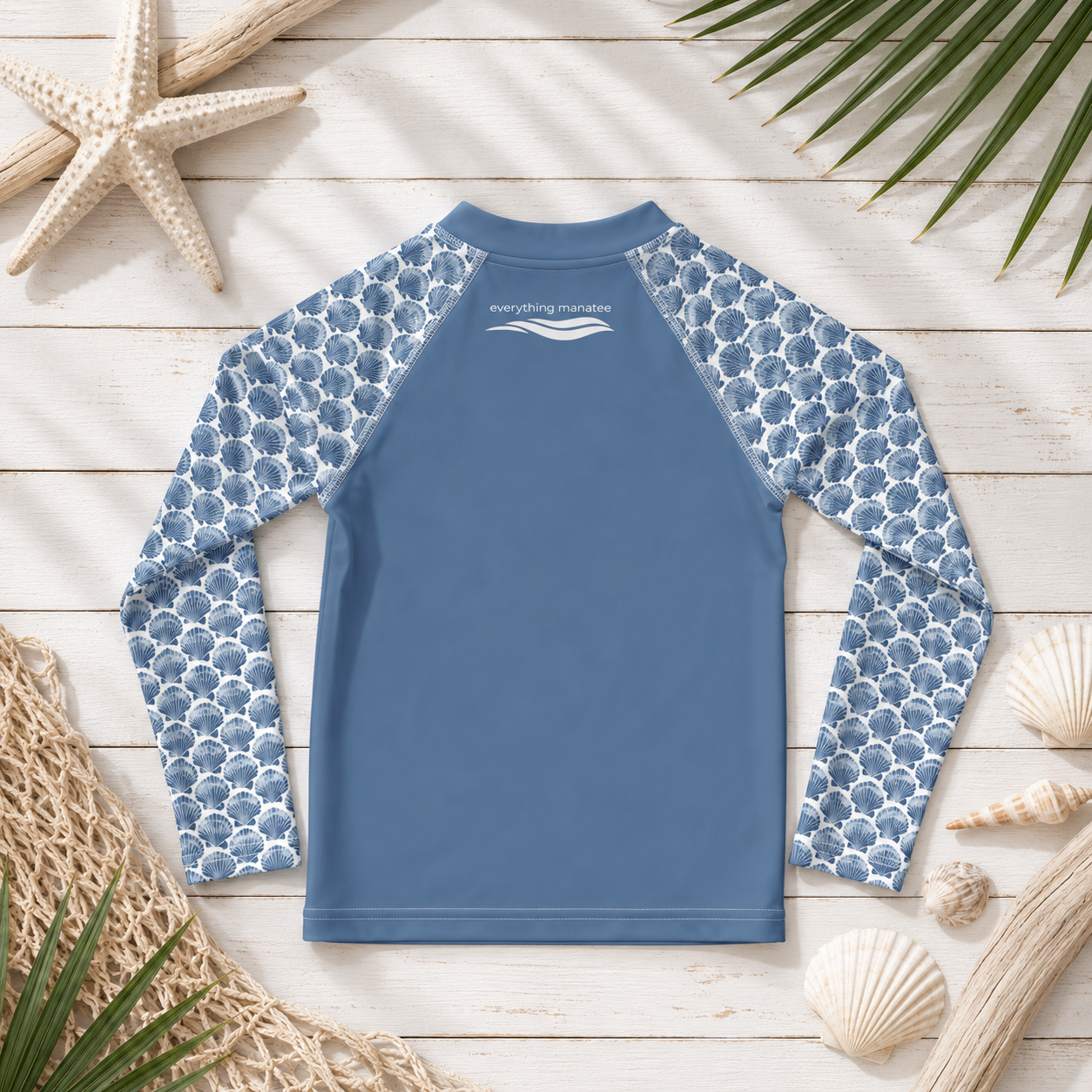 A long-sleeve kids' rash guard laid flat on a white wood plank background with beach-themed props including a starfish, palm leaves, netting, and seashells. The shirt has a solid medium blue torso and light blue scallop-patterned sleeves.