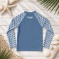 A long-sleeve kids' rash guard laid flat on a white wood plank background with beach-themed props including a starfish, palm leaves, netting, and seashells. The shirt has a solid medium blue torso and light blue scallop-patterned sleeves.