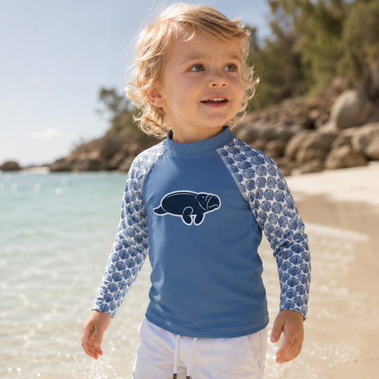 A young child standing in shallow beach water wearing a blue long-sleeve rash guard with a dark blue seal graphic on the chest and white swim shorts.