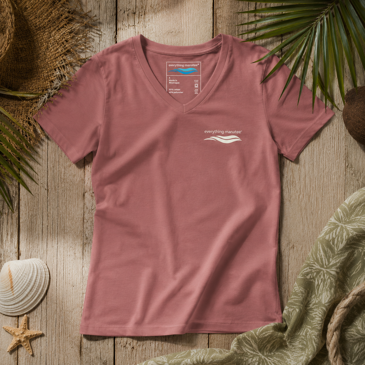 A dusty rose pink V-neck t-shirt with a small white 'everything matters' logo on the left chest, laid flat on a rustic wooden background with beach-themed props like a seashell, starfish, palm leaves, and a green towel.