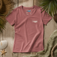 A dusty rose pink V-neck t-shirt with a small white 'everything matters' logo on the left chest, laid flat on a rustic wooden background with beach-themed props like a seashell, starfish, palm leaves, and a green towel.