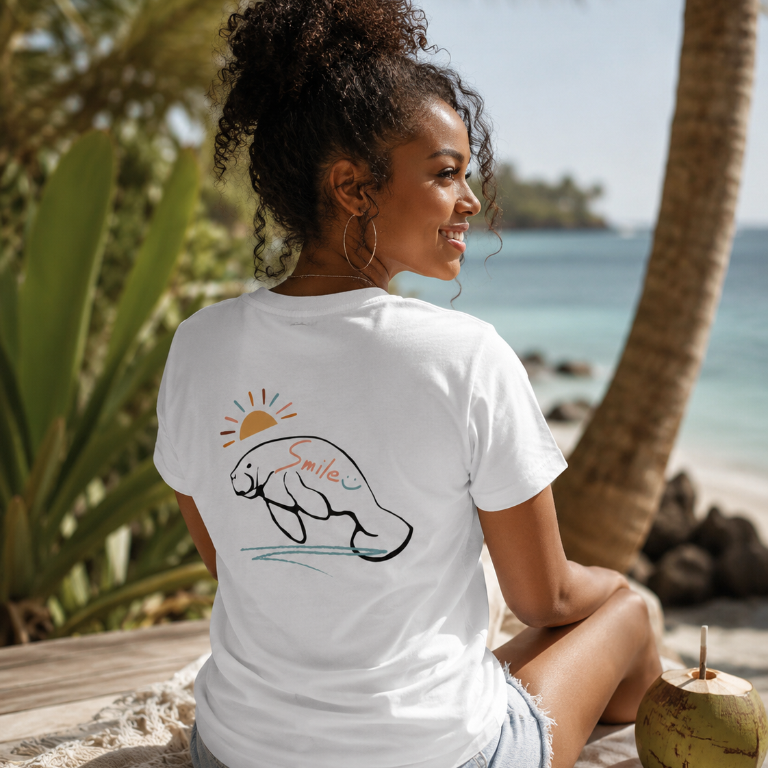 A woman sitting on a beach towel on a tropical beach, wearing a white t-shirt with a graphic of a manatee and the word 'Smile' on the back.