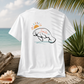 A white short-sleeve t-shirt is shown from the back, featuring a graphic of a smiling manatee under a rising sun with the word 'Smile' written across it. The shirt is displayed in a beach-themed setting with a wooden dock, seashells, a potted plant, and the ocean in the background.