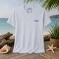 A white short-sleeve crewneck t-shirt with a small blue wave graphic and 'everything matters' text on the left chest, displayed on a beach-themed background with driftwood, seashells, a starfish, and a potted plant.
