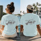 Two people sitting on a wooden dock outdoors, shown from the back wearing matching light aqua T-shirts with a gray manatee graphic and the words 'COASTAL LIFE'.