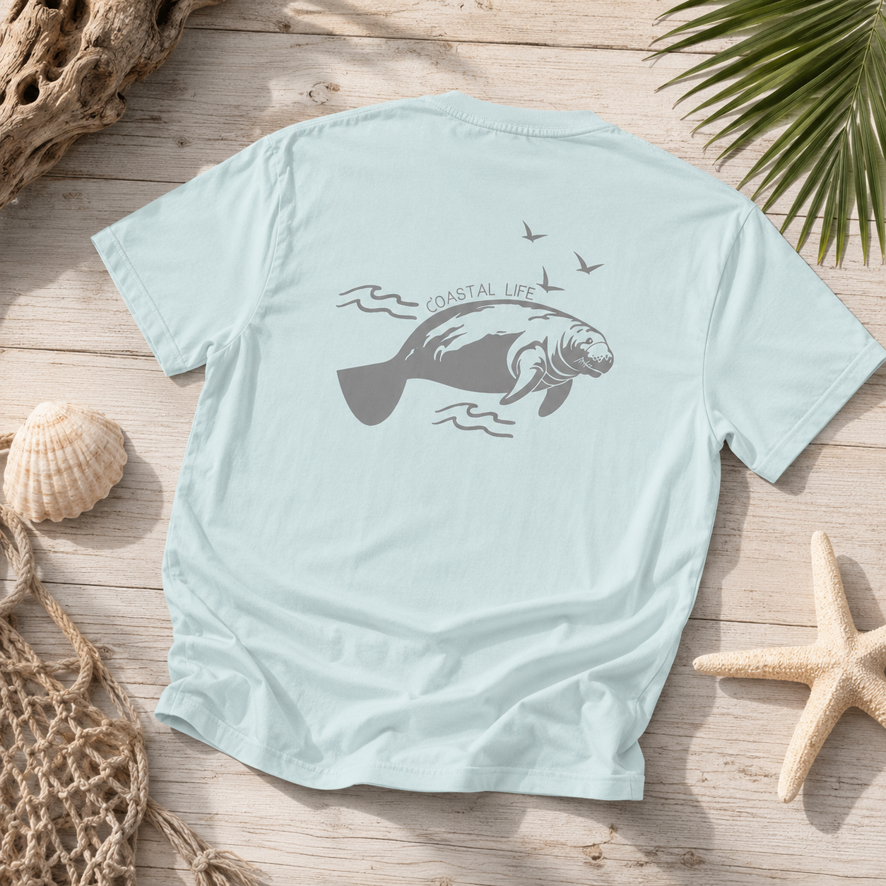 A light blue short-sleeve t-shirt with a gray manatee graphic and the words 'COASTAL LIFE' on the back, styled in a flat lay on a wooden surface with seashells, a starfish, and palm leaves.