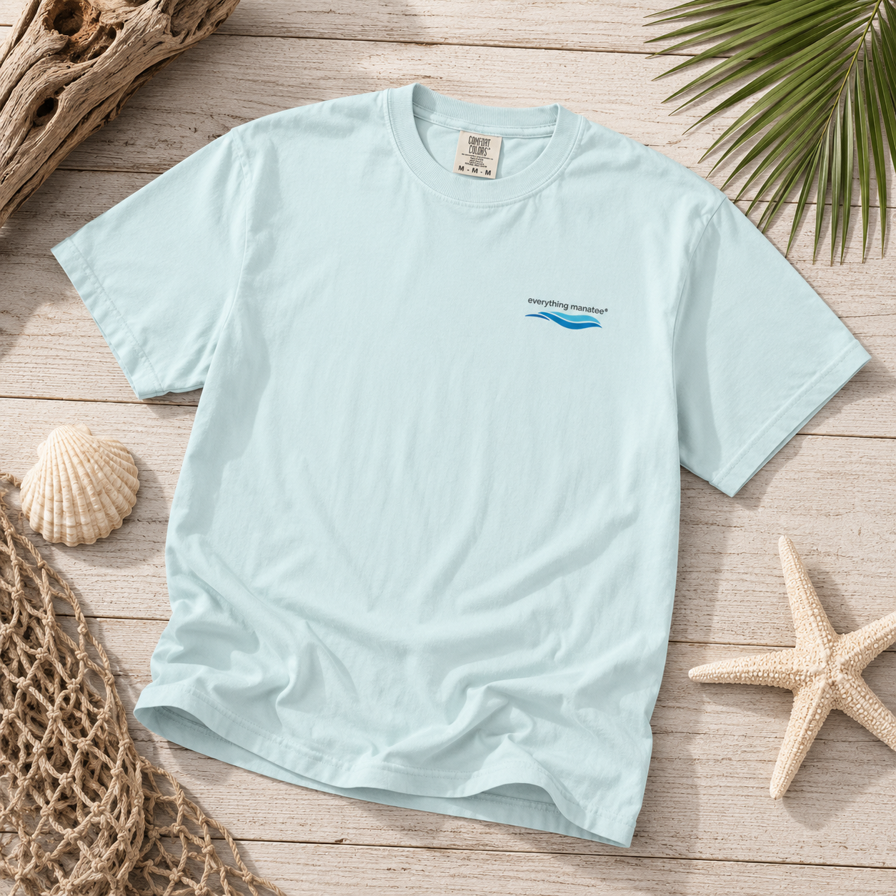 A light blue crewneck t-shirt with a small 'everything natural' wave logo on the left chest, styled in a flat lay on a wooden surface with seashells, a starfish, a net, and driftwood.