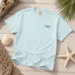 A light blue crewneck t-shirt with a small 'everything natural' wave logo on the left chest, styled in a flat lay on a wooden surface with seashells, a starfish, a net, and driftwood.