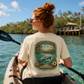 A woman in a kayak seen from behind, wearing a cream-colored t-shirt with a large graphic print of manatees and the text 'DRIFT & DREAM' on the back. She is paddling on turquoise water with tropical islands in the background.