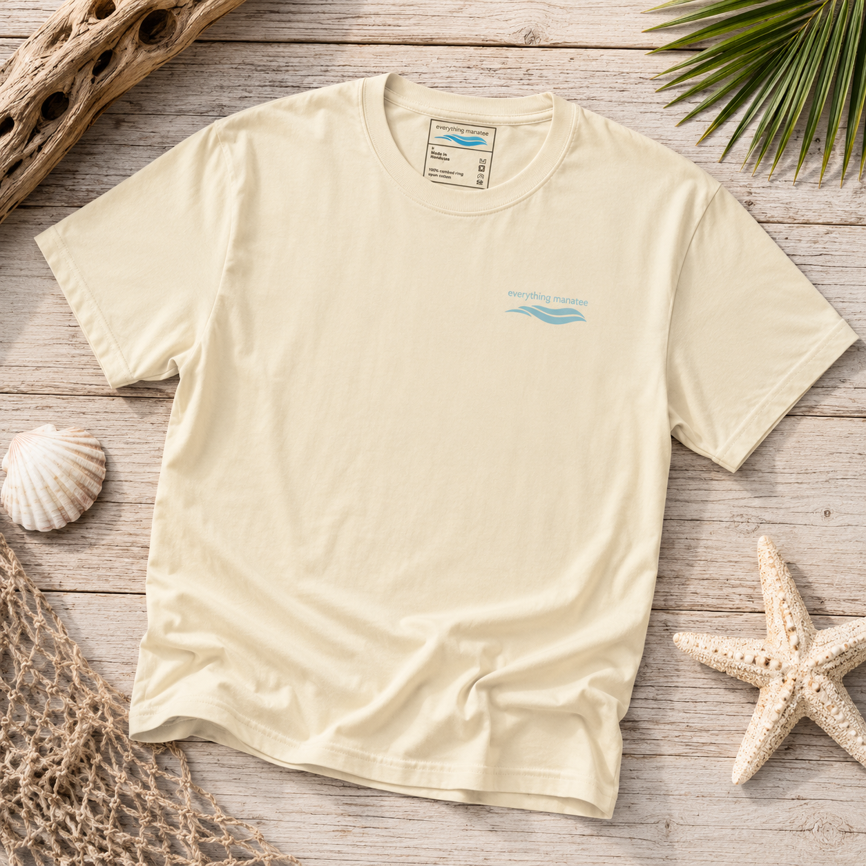 A cream-colored short-sleeve t-shirt with a small light blue wave graphic and 'Beachside Apparel' text on the left chest, laid flat on a rustic wooden surface surrounded by beach-themed props like a starfish, seashell, driftwood, and palm leaves.
