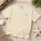 A cream-colored short-sleeve t-shirt with a small light blue wave graphic and 'Beachside Apparel' text on the left chest, laid flat on a rustic wooden surface surrounded by beach-themed props like a starfish, seashell, driftwood, and palm leaves.