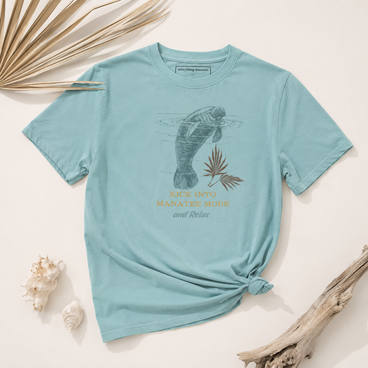 A light blue graphic t-shirt featuring a manatee illustration and the text 'RICK INTO MANATEE MODE and Relax' in gold and dark blue, styled in a flat lay with seashells, dried palm leaves, and driftwood on a cream background.
