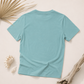 A light blue short-sleeve crewneck t-shirt laid flat on a light beige background, styled with seashells, dried palm leaves, and driftwood.