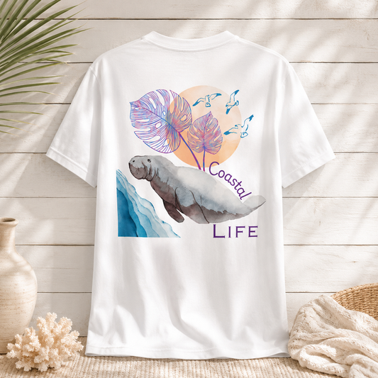 A white short-sleeve t-shirt with a colorful graphic print of a manatee, purple monstera leaves, blue waves, and the text 'Coastal LIFE', styled in a beach-themed setting with a wooden background, palm leaves, and a vase.