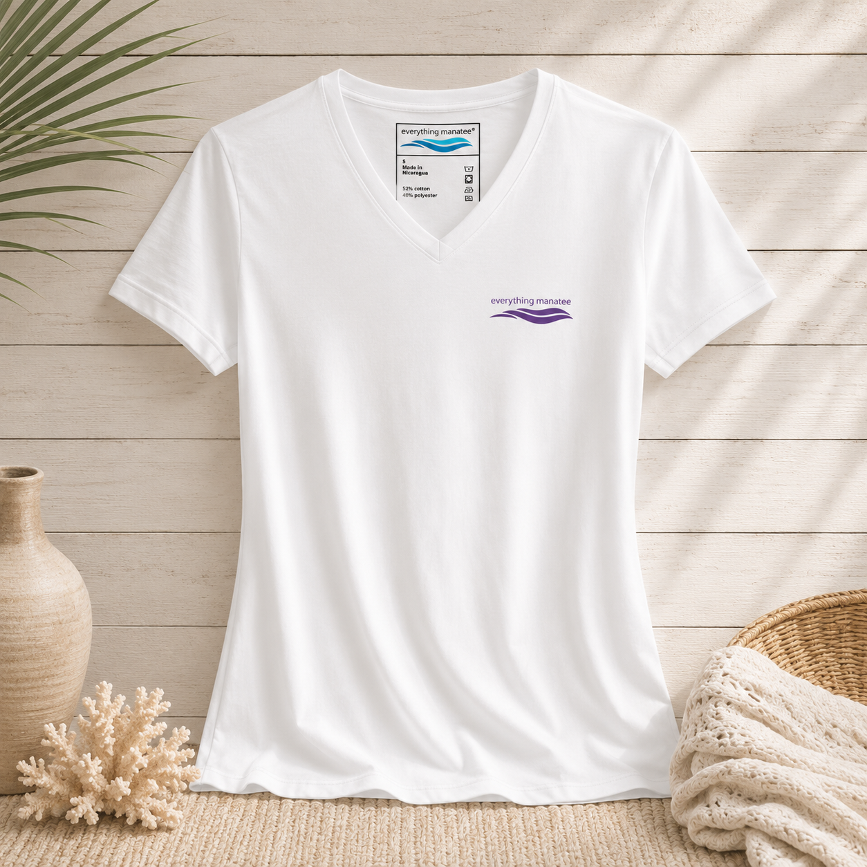 A white short-sleeve V-neck t-shirt with a small purple 'everything standard' logo on the left chest, styled in a flat-lay arrangement with a beige wood plank background, a ceramic vase, coral, and a woven blanket.