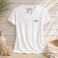 A white short-sleeve V-neck t-shirt with a small purple 'everything standard' logo on the left chest, styled in a flat-lay arrangement with a beige wood plank background, a ceramic vase, coral, and a woven blanket.