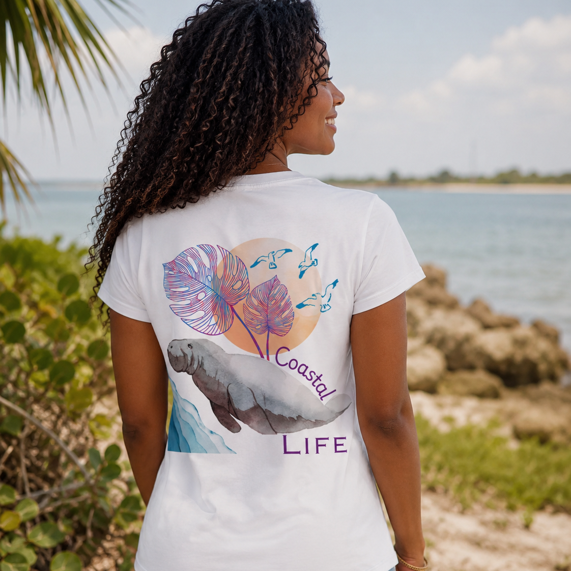 A woman standing on a beach, viewed from behind, wearing a white graphic t-shirt with a colorful print of tropical leaves, a manatee, and the words 'Coastal LIFE'.