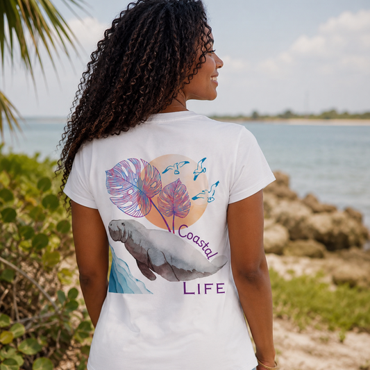 A woman standing on a beach, viewed from behind, wearing a white graphic t-shirt with a colorful print of tropical leaves, a manatee, and the words 'Coastal LIFE'.