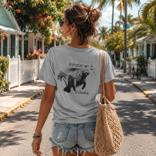 A woman wearing a light gray graphic t-shirt with a black tropical design and the words 'Inhale' and 'Exhale', paired with denim shorts and carrying a woven straw bag, walking down a sunny tropical street lined with palm trees and houses.