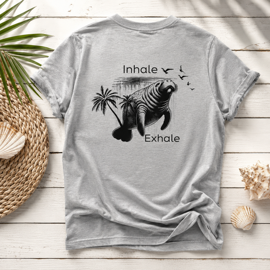 A light gray crewneck t-shirt with a black graphic of a manatee, palm trees, birds, and the words 'Inhale' and 'Exhale', laid flat on a white wood plank background with decorative seashells and a woven mat.