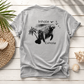 A light gray crewneck t-shirt with a black graphic of a manatee, palm trees, birds, and the words 'Inhale' and 'Exhale', laid flat on a white wood plank background with decorative seashells and a woven mat.