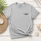 A light heather gray t-shirt with a small black 'everything marine' wave logo on the left chest, styled in a flat lay on a white wood background with a straw hat, seashells, and a palm leaf.
