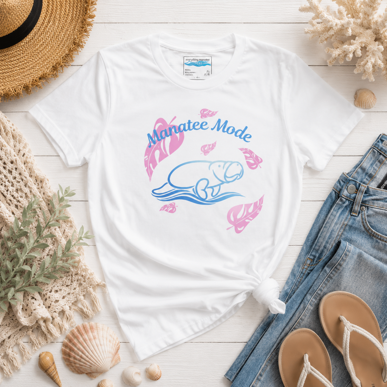 A white graphic t-shirt featuring a blue and pink manatee illustration and the text 'Manatee Mode', styled in a flat lay with jeans, sandals, a straw hat, and seashells on a white wood background.
