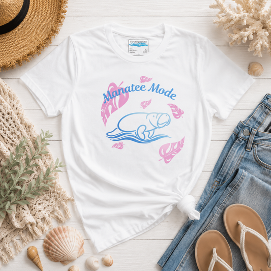 A white graphic t-shirt featuring a blue and pink manatee illustration and the text 'Manatee Mode', styled in a flat lay with jeans, sandals, a straw hat, and seashells on a white wood background.