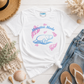 A white graphic t-shirt featuring a blue and pink manatee illustration and the text 'Manatee Mode', styled in a flat lay with jeans, sandals, a straw hat, and seashells on a white wood background.