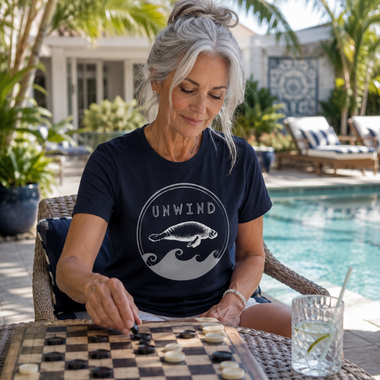 A woman with gray hair sits at a table by a swimming pool, wearing a navy blue t-shirt with a white graphic of a manatee and the word 'UNWIND'. She is playing a game of checkers on a wooden board, with a glass of limeade nearby and a resort patio in the background.