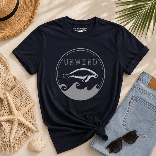 A navy blue graphic t-shirt with a gray and white manatee design and the word 'UNWIND' is styled in a flat lay on a sandy background with a straw hat, denim jeans, sunglasses, a seashell, and a woven towel.