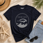 A navy blue graphic t-shirt with a gray and white manatee design and the word 'UNWIND' is styled in a flat lay on a sandy background with a straw hat, denim jeans, sunglasses, a seashell, and a woven towel.