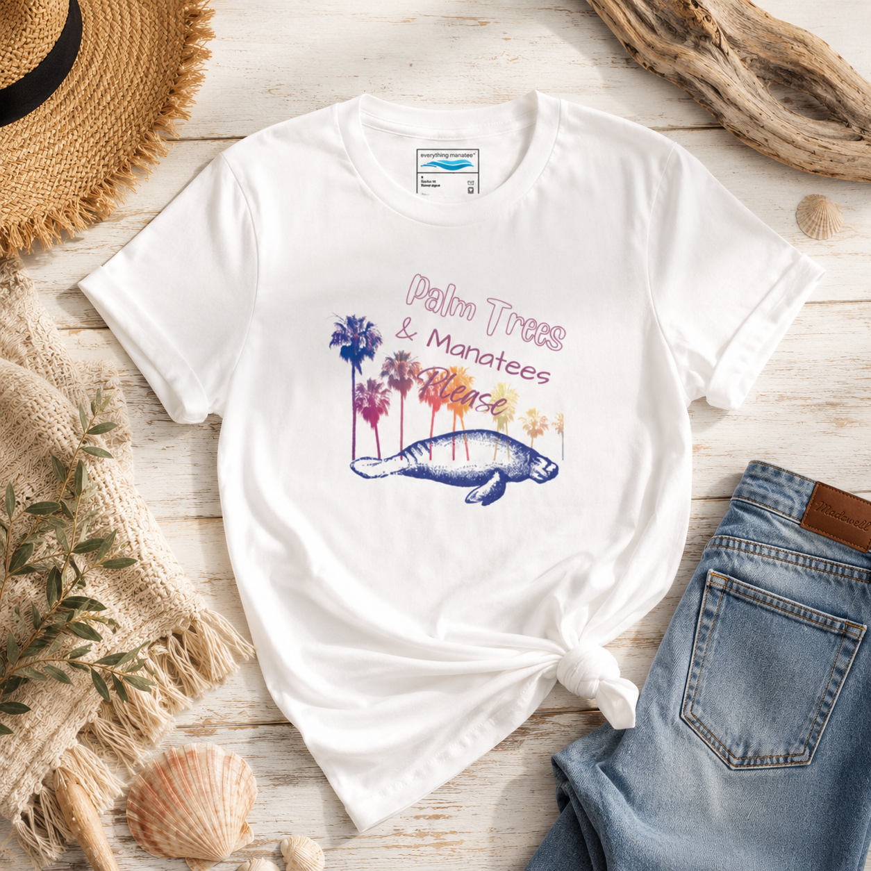 A white graphic t-shirt with a 'Palm Trees & Manatees Beach' design, styled in a flat lay with a knot at the bottom, surrounded by a straw hat, denim jeans, a woven mat, and seashells on a light wood background.