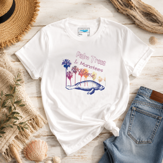 A white graphic t-shirt with a 'Palm Trees & Manatees Beach' design, styled in a flat lay with a knot at the bottom, surrounded by a straw hat, denim jeans, a woven mat, and seashells on a light wood background.