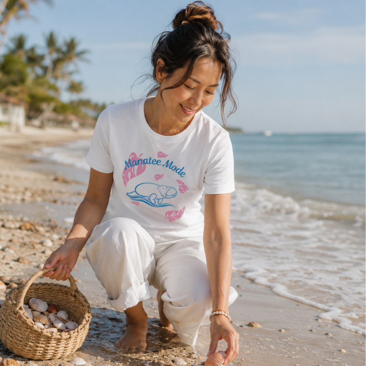 A woman crouches on a sandy beach near the water, wearing a white graphic T-shirt that reads "Mangrove Mode" with a pink and blue illustration, paired with white pants. She holds a small woven basket on the beach.
