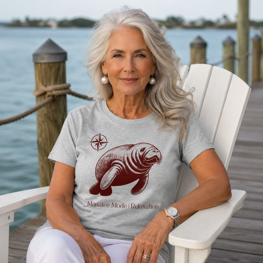 A woman with long gray hair sits in a white Adirondack chair on a wooden dock wearing a light gray t-shirt with a maroon seal graphic and the text 'Manatee Mode | Relaxation'.