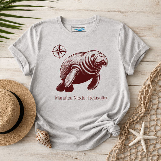 A light heather gray t-shirt with a dark red graphic of a sea otter, a compass rose, and the text 'Manatee Mode | Relaxation'. The shirt is styled in a flat-lay on a light wood background with a straw hat, a seashell, a starfish, and a net.
