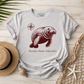 A light heather gray t-shirt with a dark red graphic of a sea otter, a compass rose, and the text 'Manatee Mode | Relaxation'. The shirt is styled in a flat-lay on a light wood background with a straw hat, a seashell, a starfish, and a net.