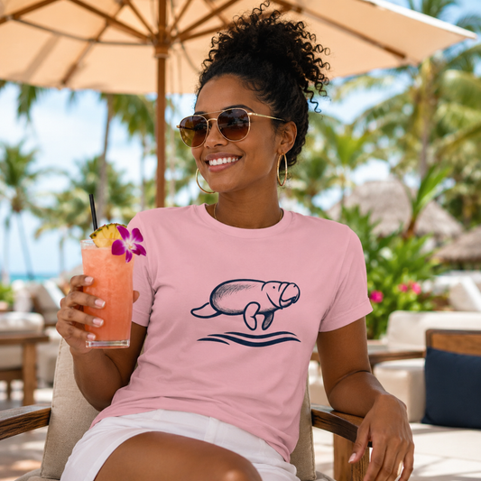 A woman wearing a light pink crewneck t-shirt with a navy blue manatee graphic, sitting in a beach lounge chair under a patio umbrella and holding a pink cocktail.
