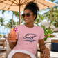 A woman wearing a light pink crewneck t-shirt with a navy blue manatee graphic, sitting in a beach lounge chair under a patio umbrella and holding a pink cocktail.
