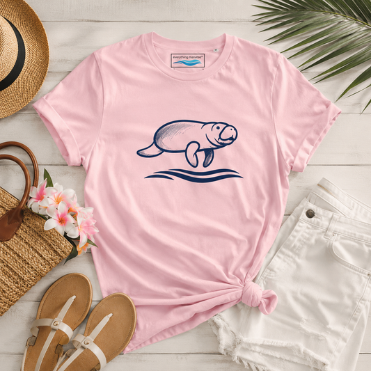 A pink short-sleeve t-shirt with a navy blue manatee graphic, styled in a flat lay with white shorts, a straw hat, sandals, flowers, and a woven bag on a white wood background.