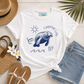 A white graphic t-shirt featuring a navy blue manatee illustration and the words 'coastal life' with beach-themed props including denim shorts, a straw hat, sandals, and flowers on a light wood background.