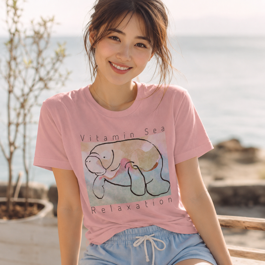 A woman wearing a light pink graphic t-shirt with a turtle illustration and the text 'Vitamin Sea Relaxation', paired with light blue shorts, set against a blurred outdoor background of water and rocks.