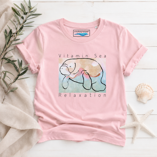 A light pink short-sleeve t-shirt with a graphic of a manatee and the text 'Vitamin Sea Relaxation' on the front. The shirt is styled in a flat-lay arrangement on a white wood background with a beige linen cloth, green leaves, a seashell, and a starfish.