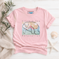 A light pink short-sleeve t-shirt with a graphic of a manatee and the text 'Vitamin Sea Relaxation' on the front. The shirt is styled in a flat-lay arrangement on a white wood background with a beige linen cloth, green leaves, a seashell, and a starfish.