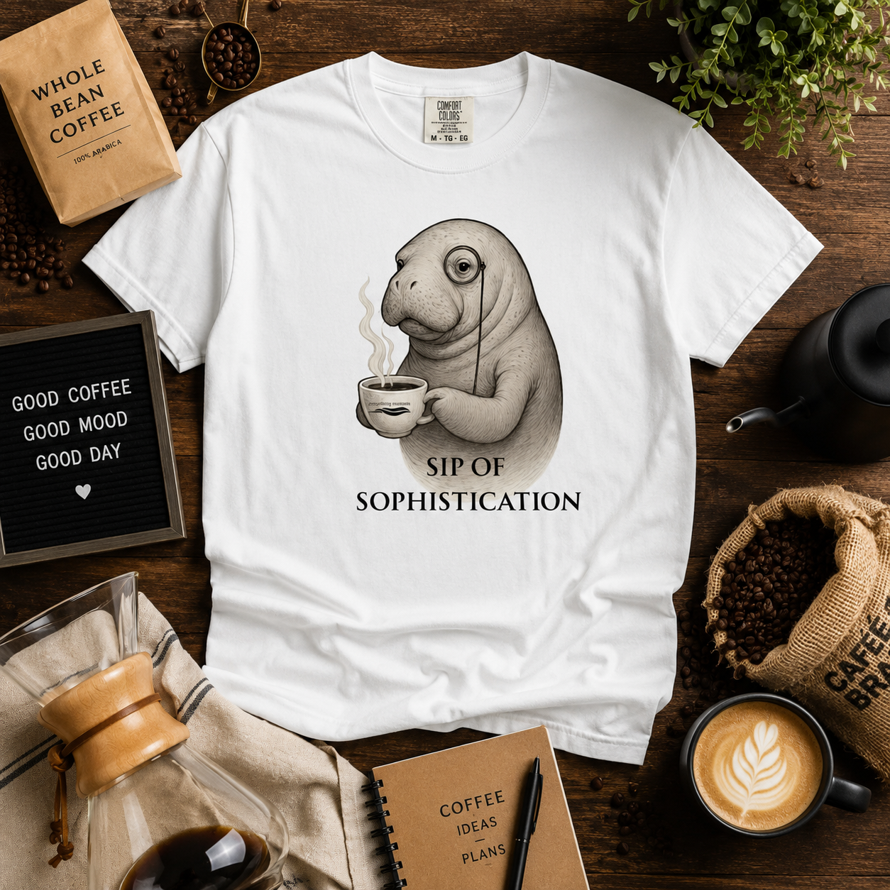 A white short-sleeve graphic T-shirt laid flat on a rustic wooden surface, featuring a sepia illustration of a seal holding a coffee cup with the text 'SIP OF SOPHISTICATION.' The shirt is styled with coffee-themed props including a coffee bag, coffee beans, a coffee maker, a cup of latte, a notebook, and a letter board.