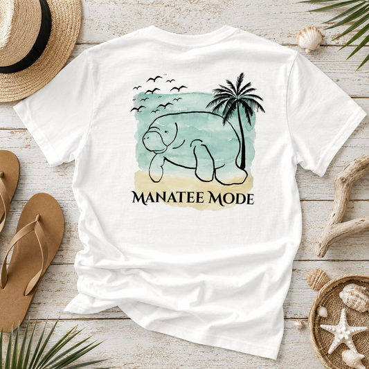 A white short-sleeve graphic T-shirt laid flat on a whitewashed wood surface, surrounded by straw hat, sandals, driftwood, and seashells. The shirt features a beach-style illustration of a manatee with a palm tree and the text 'MANATEE MODE'.