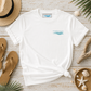 A white short-sleeve t-shirt with a small light blue chest graphic reading 'everything marine' with wave lines, styled in a flat lay on a whitewashed wooden surface with straw hat, sandals, shells, driftwood, and palm leaves.