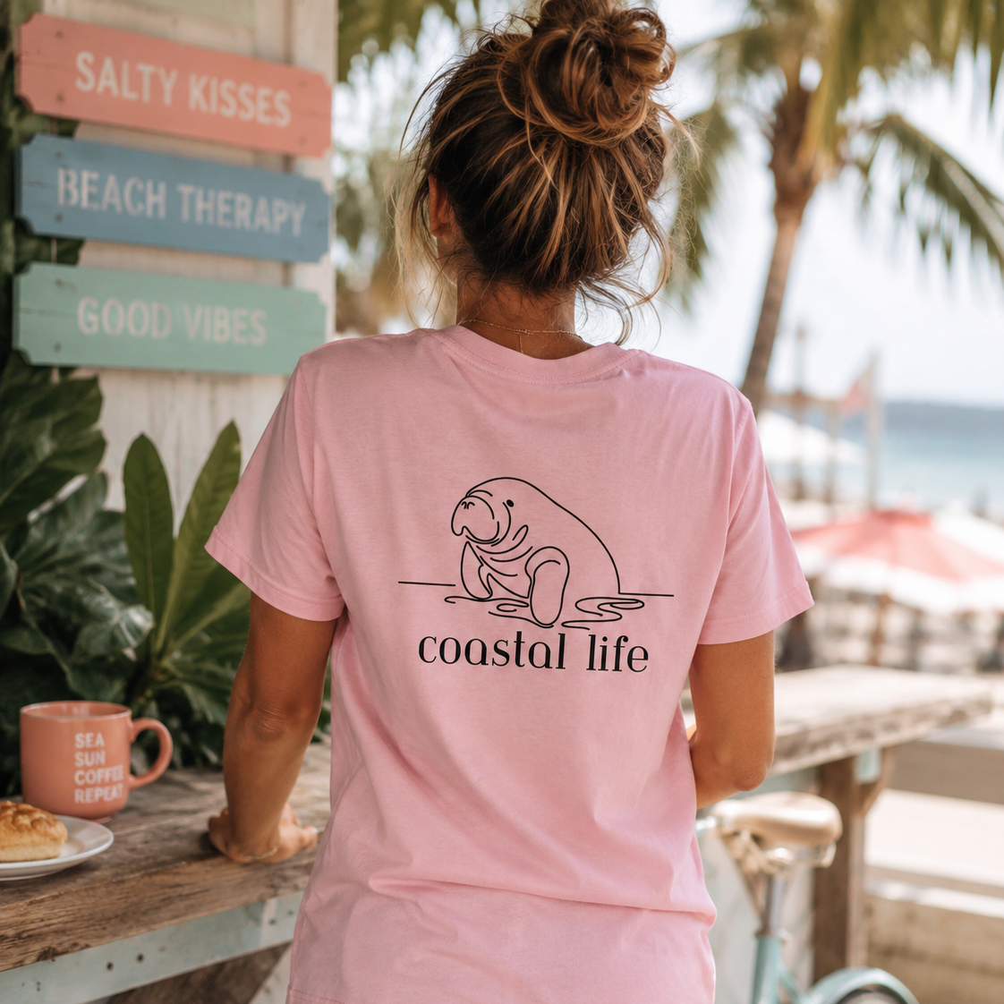 A woman viewed from behind wearing a light pink t-shirt with a black line art graphic of a manatee and the text 'coastal life' printed on the back. She is standing outdoors at a beach cafe next to a light blue bicycle, with a wooden sign reading 'SALTY KISSES' in the background.