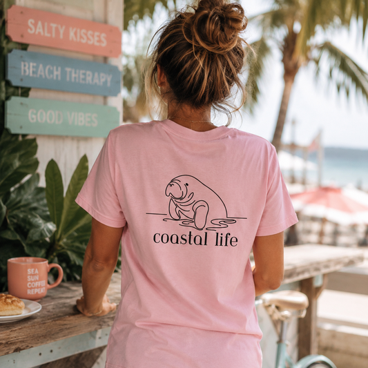 A woman viewed from behind wearing a light pink t-shirt with a black line art graphic of a manatee and the text 'coastal life' printed on the back. She is standing outdoors at a beach cafe next to a light blue bicycle, with a wooden sign reading 'SALTY KISSES' in the background.